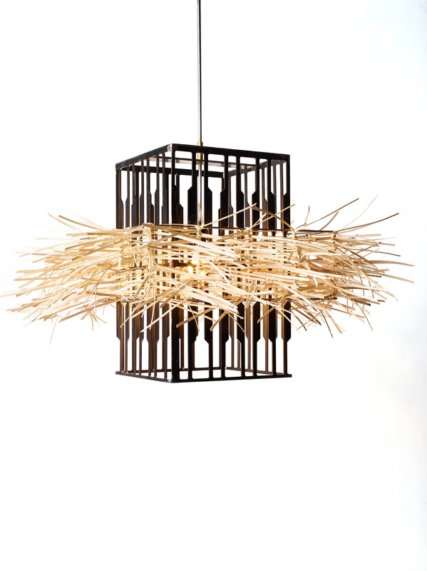 Suspended artistic lighting created by Lucy Slivinski with a vertical sitting rectangular prism made from scrap metal punch outs and thin wooden reed woven into the center portion, diffusing the single bulb inside.
