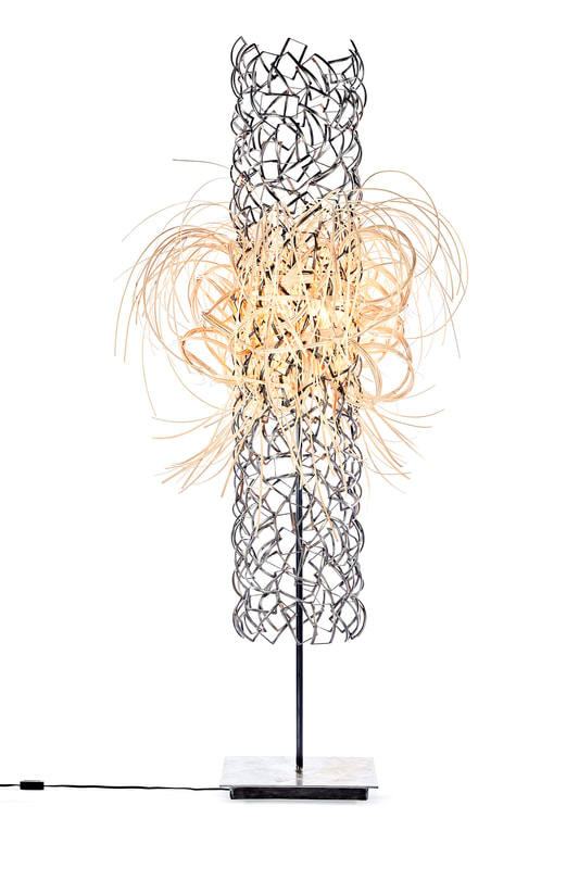 Tall sculptural floor light made from scrap metal pieces welded into a long cylinder form from which various wooden reed is woven into the center, flaring out in all directions, diffusing the single light bulb. Created by Lucy Slivinski.