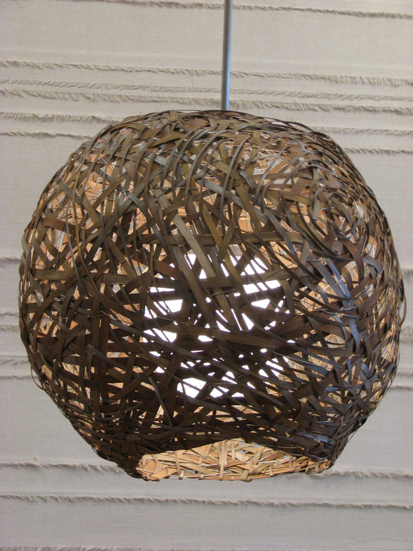 Suspended woven light made from wooden reed and wire hand woven into a spherical shape encasing a single glass globe while open at the bottom. Created by Lucy Slivinski.