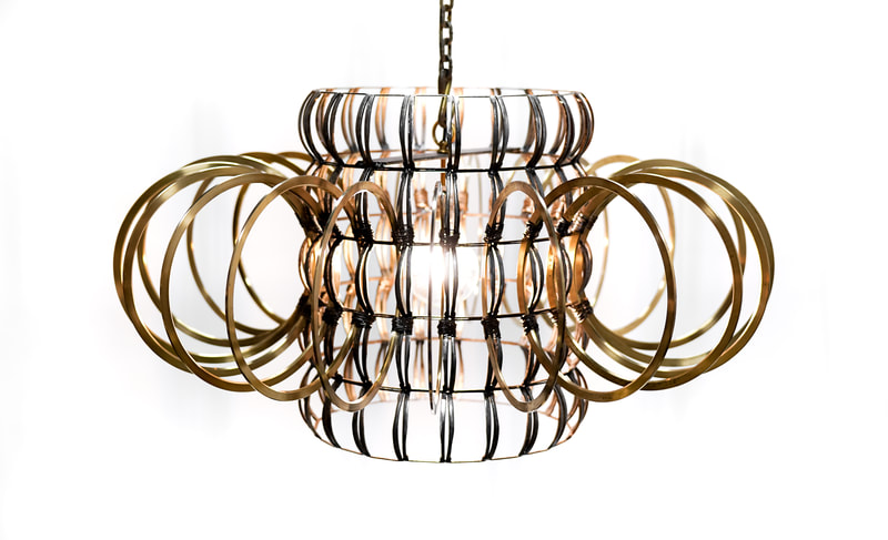 Suspended artistic lighting created by Lucy Slivinski using scrap metal to form a vertical standing cylinder with brass rings encircling it perpendicularly. Single bulb inside.