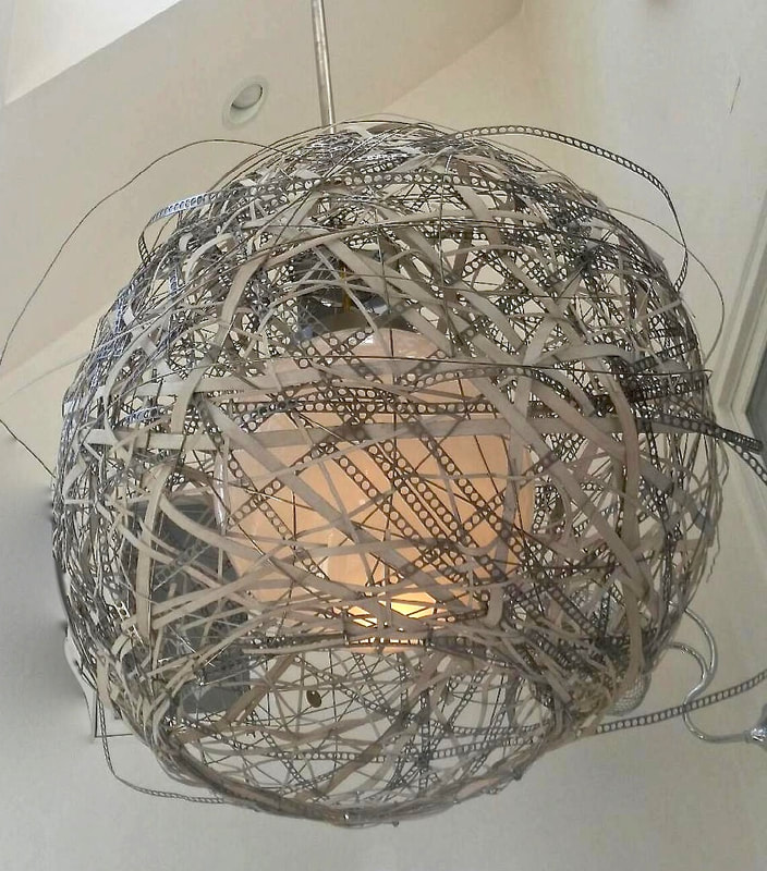 Suspended woven light made from mostly stainless steel wire and some wooden reed hand woven into a spherical shape encasing a single glass globe while open at the bottom. Created by Lucy Slivinski.