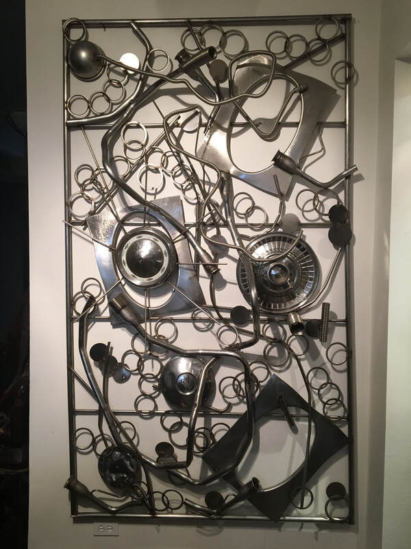 Wall mounted abstract metal sculpture made from salvaged stainless steel rings, various shaped cut outs and car hubcaps mounted onto metal frame. Created by Lucy Slivinski. 