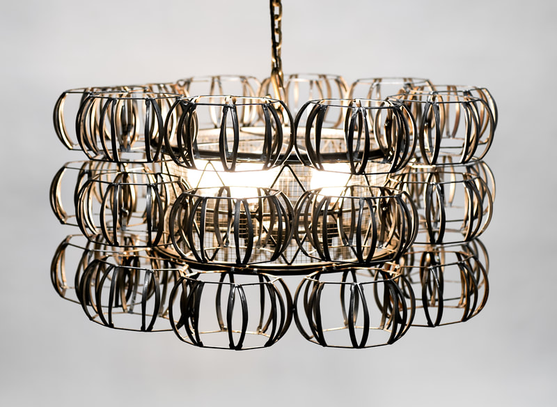 Large suspended artistic lighting created by Lucy Slivinski with a central cylinder encasing three bulbs and three rows of numerous metal spherical forms encircling the central form.