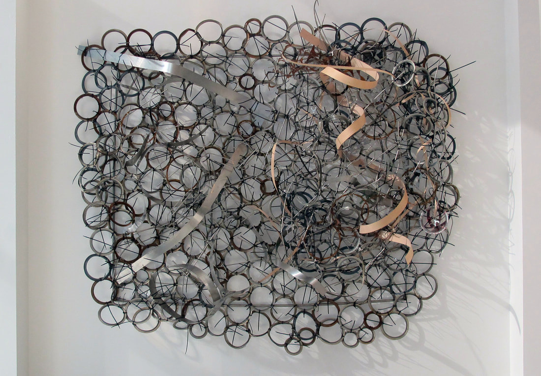Large wall mounted abstract sculpture made from found objects.  Sheet of paint can rings, black zip ties and strips of wood and metal woven in. Created by Lucy Slivinski. 