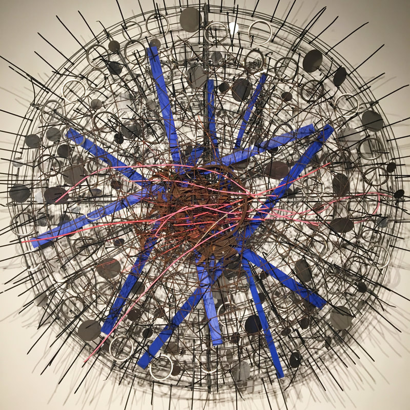 Large wall mounted abstract sculpture made from found objects.  Large metal fan grate with black zip ties, blue plastic strips and metal discs woven in. Created by Lucy Slivinski. 