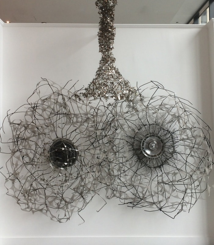 Two large wall mounted abstract sculptures made from salvage metal and found objects positioned side by side with sculptural lighting hanging over head and center to the wall pieces.