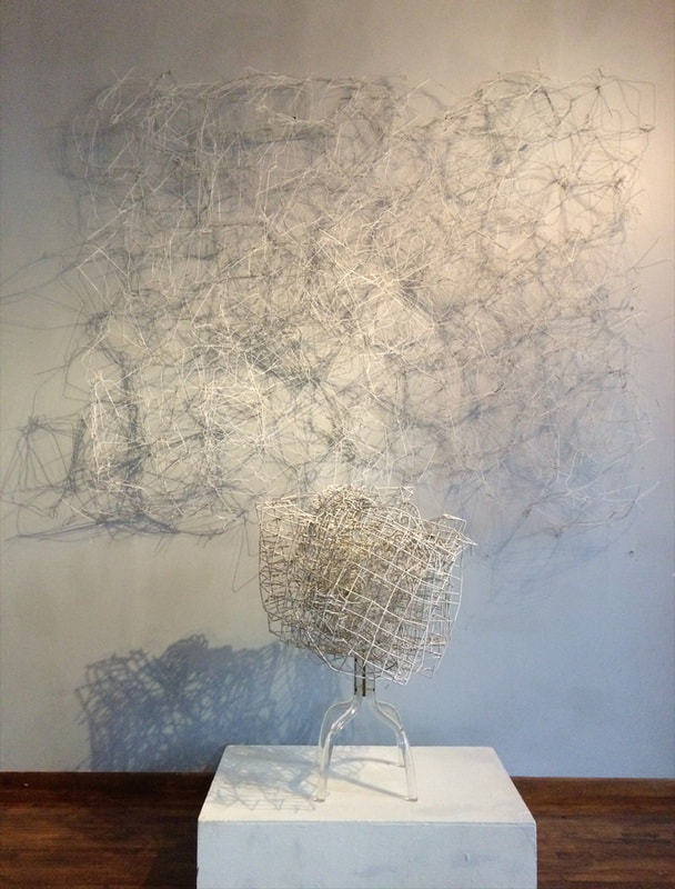 Large abstract sculpture made from found objects.  White coat hangers woven into a sheet flat against the wall and a mangled dish washer rack mounted on the ground in front. Created by Lucy Slivinski. 